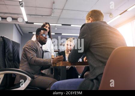 African male wearing tuxedo sit speaking with caucasian man while others listening to them. Teamwork in business center Stock Photo