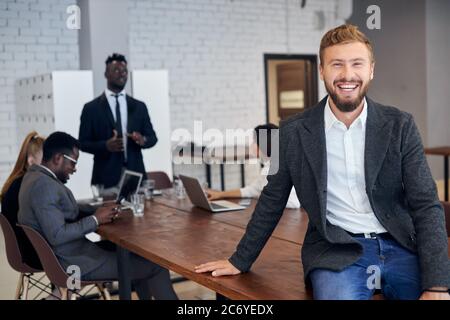 Handsome young man in tuxedo and eyeglasses using smartphone in workplace while his business colleagues having conversation in background Stock Photo