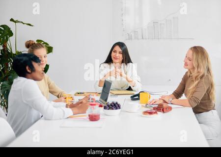 Young business ladies interacting during coffee break at business conference in white office, fruits and cup of tea on table. women colleague gathered Stock Photo