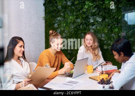 Successful teamwork of young diverse business people, female business partners co-working together using laptop. White modern office with green wall Stock Photo