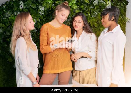 attfour attractive diverse women gathered to discuss business ideas, use mobile phone during break time at work place Stock Photo
