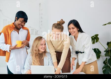 Caucasian and african women, business ladies gathered in modern white office to discuss business ideas using laptop, teamwork of beautiful ladies wear Stock Photo