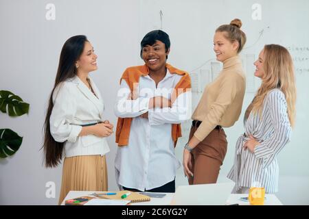 Portrait of successful team of four diverse business ladies dressed in casual wear and stand smiling, discussing, look at each other. light modern bus Stock Photo