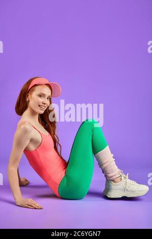 red-haired woman rests after aerobics workout, sits on floor pulled up legs, drese in pink leotard, green tight leggins and white training shoes, look Stock Photo