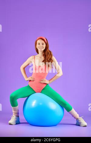sporty ginger girl who sits relaxed on blue fit ball after active cardio training, feels satisfied, full of energy and positive emotions, wears pink l