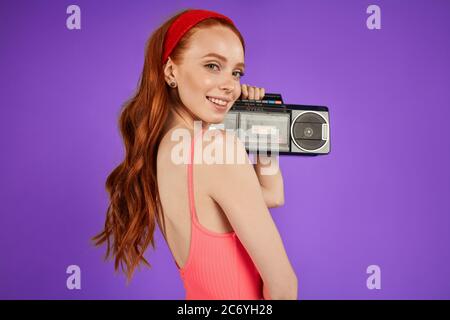 back view of motivated charming red-haired girl with green eyes and cute freckles, stands with portable audio player, looks playfully in camera with l Stock Photo