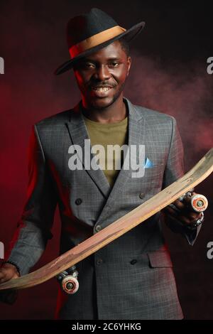 Portrait of energetic, sporty, self-assured handsome african man infashionable blazer suit holding longboard in hands, posing isolated in studio on da Stock Photo