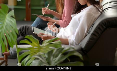 Side view of female college students relaxed sitting on black couch and using digital tablets with stylus in living room Stock Photo