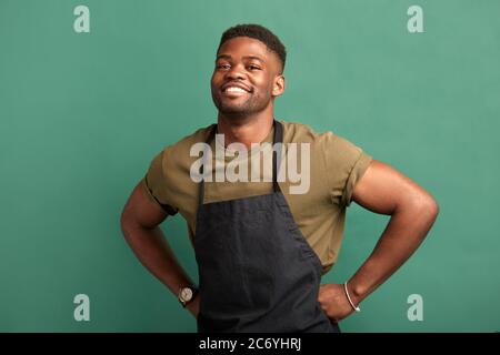 African male farmer with muscular hands and sportive body, dressed in apron smiling at camera with toothy smile over green background Stock Photo