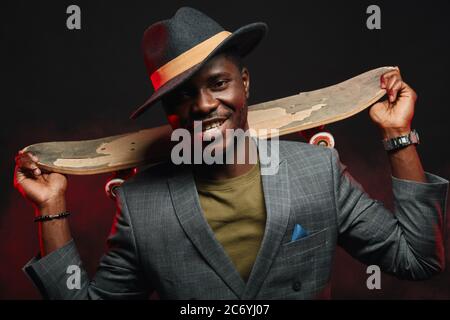 Young african man in formal jacket and hat holds skateboard in hands. Busines and Sport, Healthy lifestyle concept Stock Photo