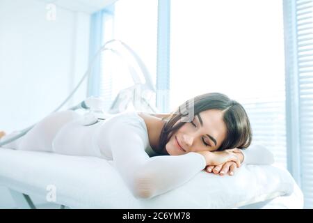 Attractive brown-haired woman in white elastic suit is lying on coach at modern medical clini , getting LPG body massage and treatment using hardware Stock Photo