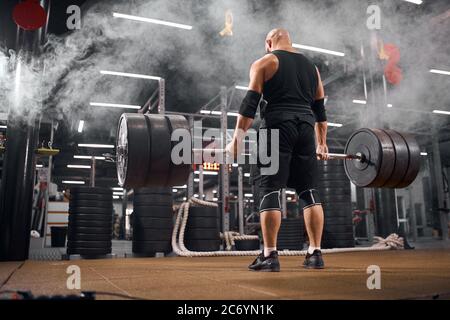 Young hairless bodybuilder keeping heavy weight barbell, standing in white smoke, trying to do deadlift, training in gym, back side view, indoor shot Stock Photo