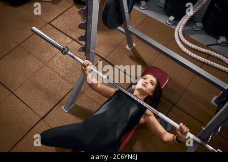Beautiful sportive woman keeping hand on iron bar lying on red bench under heavy barbell, trying to lift weight, looking up with concentrated face, ta Stock Photo