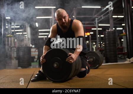 Young man with bald head, dressed in black stylish sportswear looks aside with interest, sitting on barbell, changing metal weight plate, gym atmosphe Stock Photo