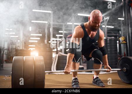Hairless, severe bodybuilder doing barbell squats in modern fitness center, brutal man practicing for training using gym equipment, white smoke backgr Stock Photo
