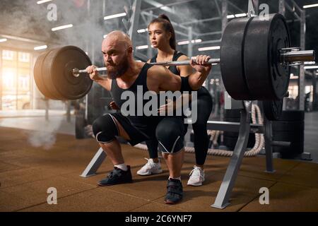 Strong brutal male powerlifter doing squats using heavy barbell, young active female trainer showing the right position, preparing for serious competi Stock Photo