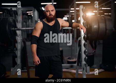 Sportive young man modeling in modern fitness club, posing near metal bar, putting hand on professional barbell, looking straight at camera, tired aft Stock Photo