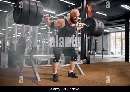 Young strong bodybuilder preparing for competition of powerlifters, doing squats using heavy metal barbell, looking away with rude face, training hard Stock Photo