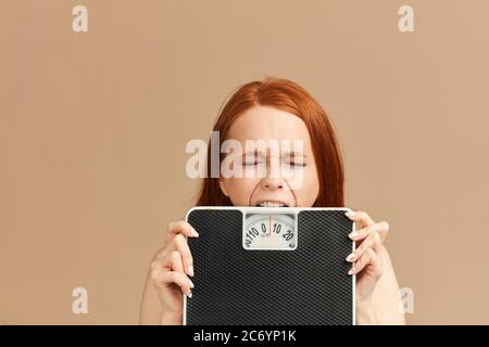 Young woman crunching mechanic scales in hands feeling sad and depressed, trying to bite scales in dispair, isolated on beige background Stock Photo