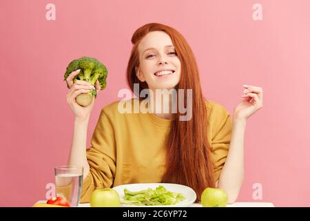 Young beautiful happy attractive female with gorgeous long ginger hair having dinner with fresh broccoli and vegetables at table, isolated over pink b Stock Photo