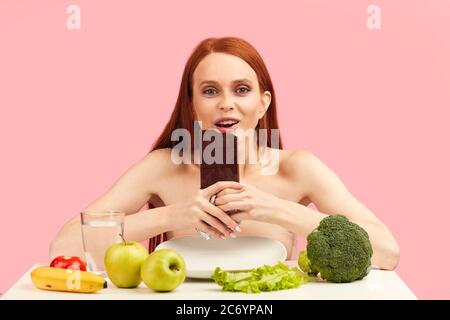 Happy young skinny woman exhausted by severe diets, holding chocolate bar, posing isolated on pink. Diet failure of emaciated redhead woman is eating Stock Photo
