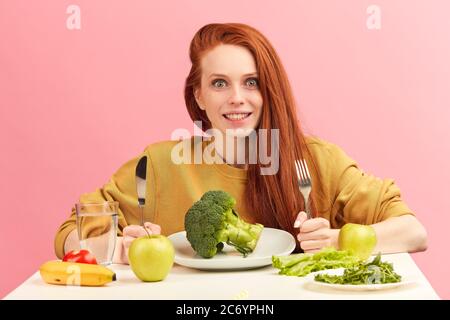 Hungry young vegg woman eating with eagerness and appetite raw green broccoli , smiling happy at camera over studio isolated pink background. Vegetabl Stock Photo