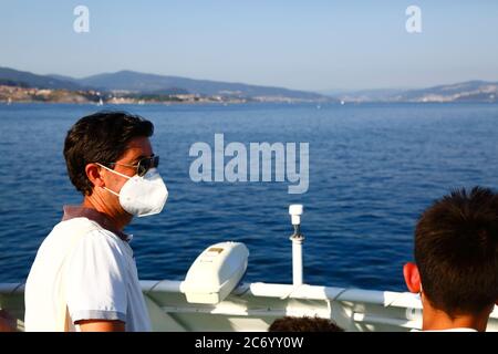 12th July 2020, Ria de Vigo, Galicia, northern Spain: A male passenger wearing a face mask and sunglasses looks at the view from the ferry between Vigo and the Cies Islands (a popular tourist destination off the coast of Galicia). Spain has relaxed travel restrictions from 21st June after a strict lockdown to control the Covid 19 coronavirus and many Spaniards are returning to the beaches. Wearing face masks is still mandatory on public transport. Stock Photo