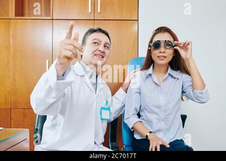 Smiling ophthalmologist testing eyesight of young female patient and asking her to read letters on testing board Stock Photo
