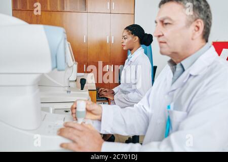 Ophthalmologist working on medical equipment when his assistant working on computer and entering patients information Stock Photo