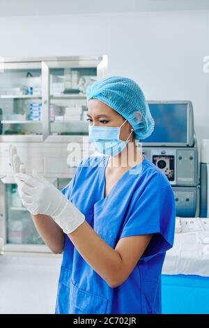 Serious young surgical assistant in scrubs and medical mask putting on sterilized surgical gloves operating room Stock Photo