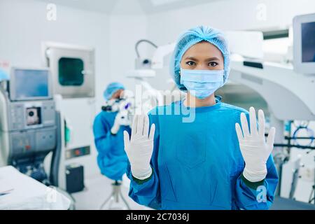 Young Asian female surgeon in scrubs and medical masks showing hands in white sterilized gloves Stock Photo