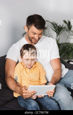 happy father with attentive son sitting on sofa and using laptop together Stock Photo
