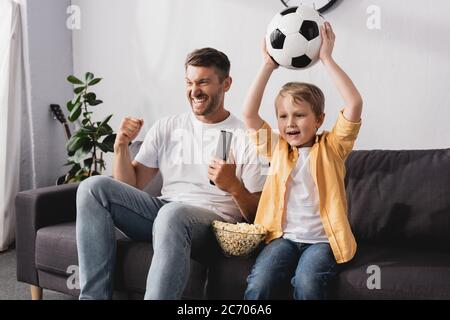excited man showing winner gesture near son holding soccer ball in raised hands while watching tv Stock Photo
