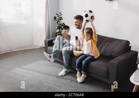 excited man showing winner gesture near son holding soccer ball in raised hands while watching tv Stock Photo