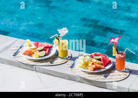 Two plates with fresh fruit and two juice with slice orange. Healthy breakfast on poolside in hotel. Plate with papaya, pineapple, watermelon, lime. Stock Photo