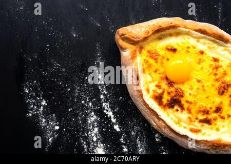 Khachapuri adjara traditional Georgian cuisine meal. Baked bread as boat with cheese and egg filling on black background, decorated with flour. Nation Stock Photo