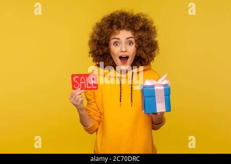 Wow, bonuses on Black Friday shopping! Portrait of surprised overjoyed curly-haired woman in urban style hoodie showing Sale inscription and gift box. Stock Photo