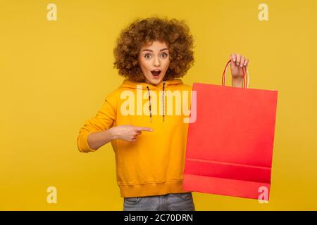 Portrait of amazed shocked curly-haired hipster girl in hoodie pointing at shopping bags with blank area, copy space on packages for advertising text. Stock Photo