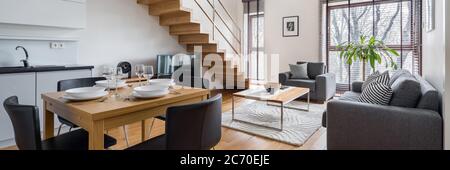 Panorama of stylish two-floor apartment with kitchen and dining area open to living room and wooden stairs to higher floor Stock Photo