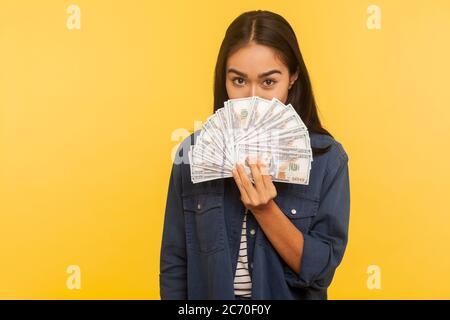 Portrait of happy brunette girl in denim shirt peeking out of dollar banknotes with playful happy look, showing money, big profit, financial savings. Stock Photo