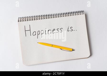 Iscription HAPPINESS IS in the notebook on white background. Top view of notebook and pen. Stock Photo