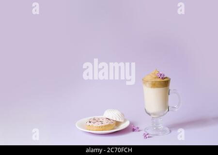 Beautiful dalgona drink a frothy coffee in a transparent mug and flowers lilac to purple Il purple background. Sweets on a plate: Donat and tender mar Stock Photo