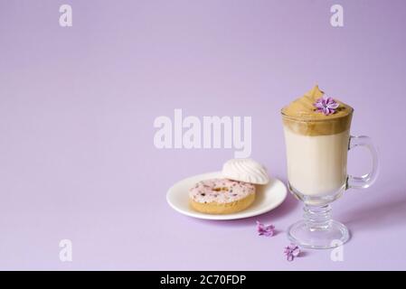 Beautiful dalgona drink a frothy coffee in a transparent mug and flowers of lilac on a purple background. Sweets on a plate: Donut and tender marshmal Stock Photo