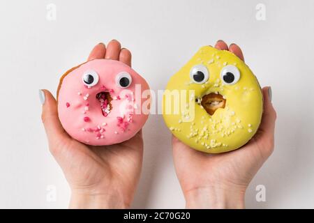 Female hands holding two donuts with funny faces and huge eyes. Yellow and pink doughnut on white background. Stock Photo