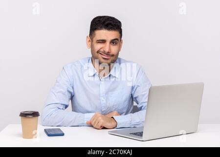 Handsome elegant businessman sitting office workplace with laptop on desk, winking playfully at camera and flirting, blinking eye having interesting i Stock Photo
