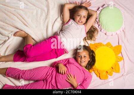 Kids in pink pajamas have fun, copy space. Children with smiling faces lie close on light pink blanket background. Childhood and happiness concept. Schoolgirls have pajama party with funny pillows. Stock Photo