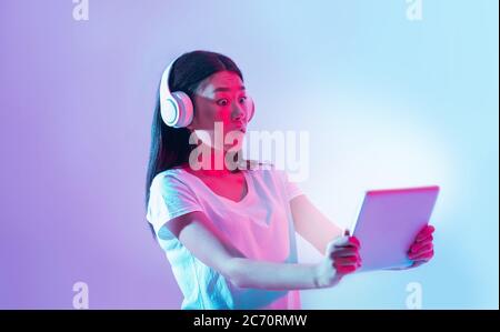Modern devices are surprised. Shocked asian girl in white headphones looks at tablet with luminous screen Stock Photo