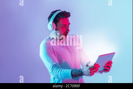 Amazing surprise online. Shocked man with open mouth in headphones holds tablet neon light Stock Photo