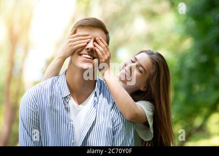 Affectionate girlfriend closing her boyfriend's eyes, playing guess who game in park Stock Photo