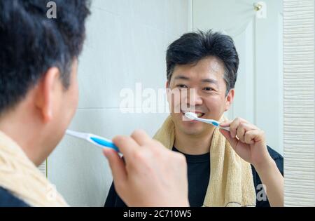A middle-aged Asian man who wears a towel around his neck and brushing teeth in the bathroom. Stock Photo